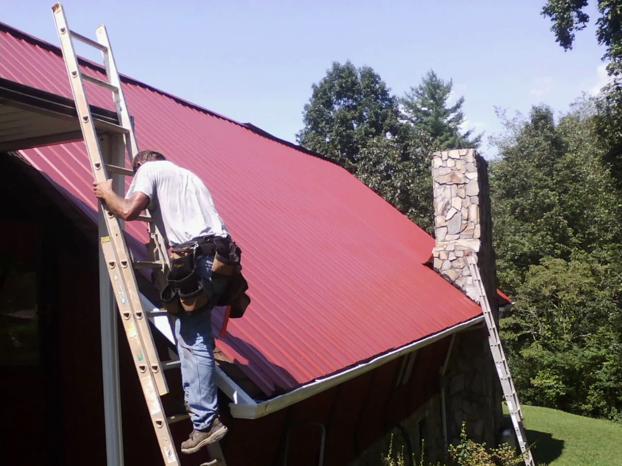 Roofer on a ladder beside a red metal roof and stone chimney — Soffit & Fascia Repair by Roofing Brothers in Ellerslie